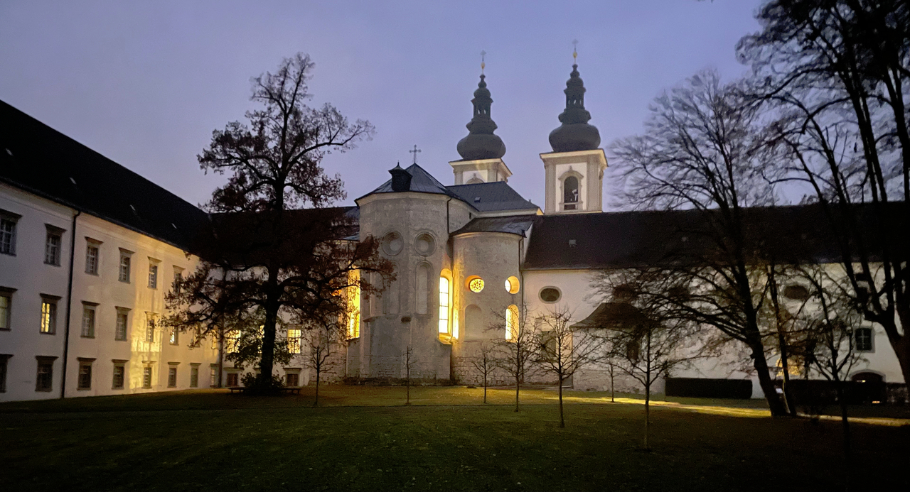 Die Stiftskirche aus Blickrichtung des Innenhofes der Mönche. Es ist Nebel und Abenddämmerung. Die Fenster der Kirche strahlen warmes Licht aus.