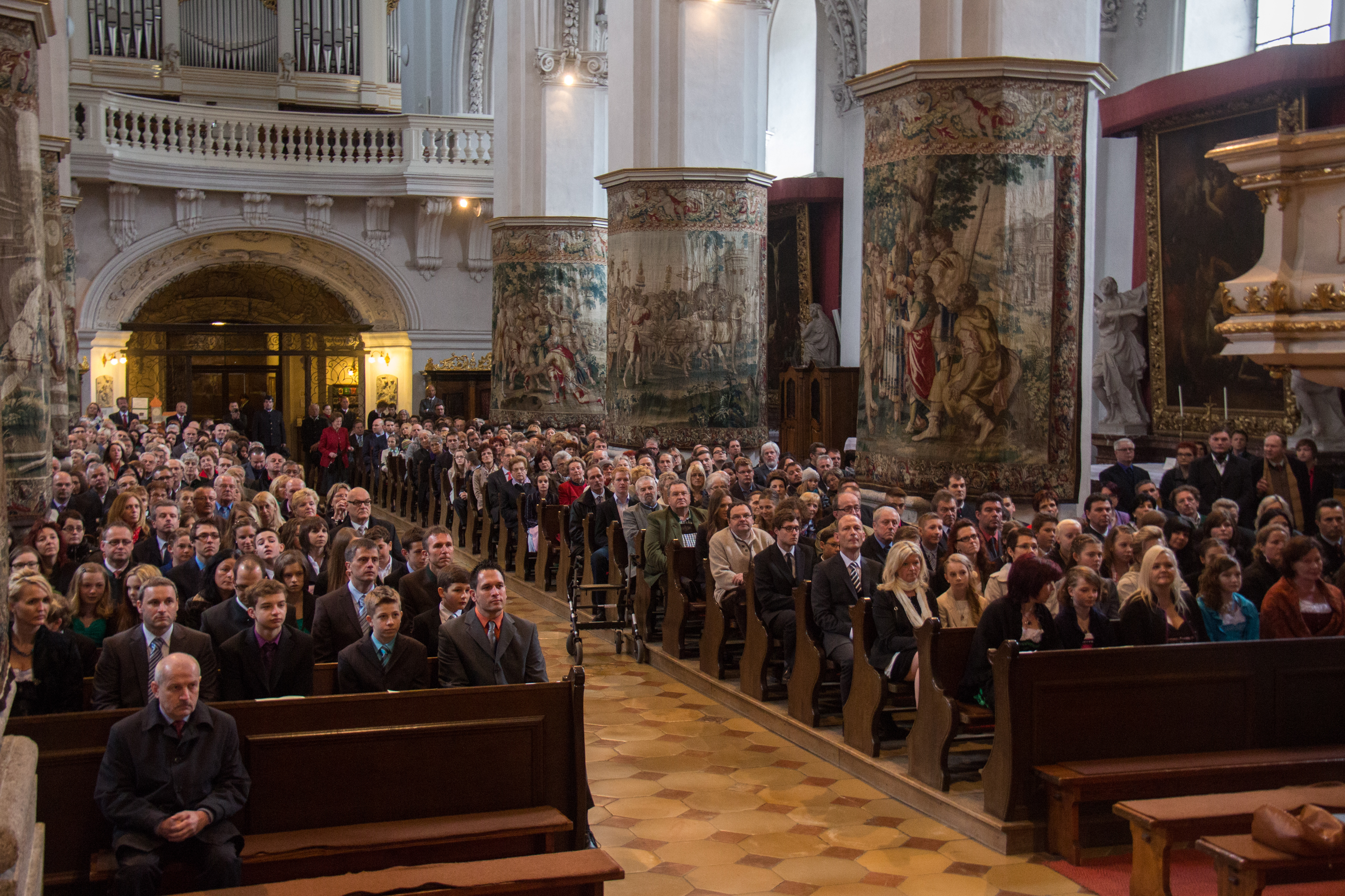 Das Mittelschiff der Stiftskirche ist voll mit Gottesdienstbesucher, aufwendig bestickte Teppiche verzieren die dicken Säulen der Stiftskirche 