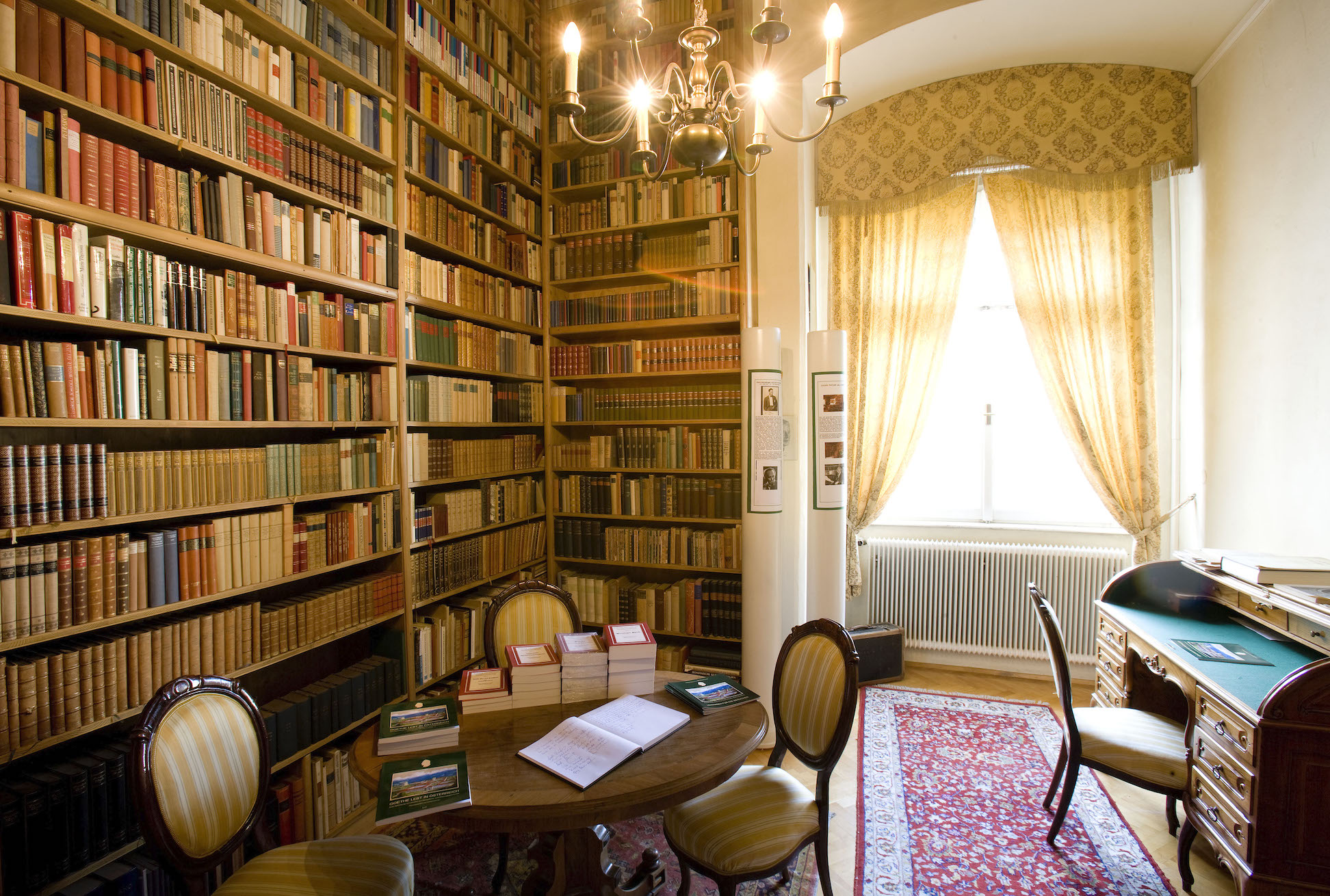 Zimmer mit einer Wand voll mit Büchern, einem Kronleuchter, Sitzgelegenheiten zum Besprechen und ein Schreibtisch 