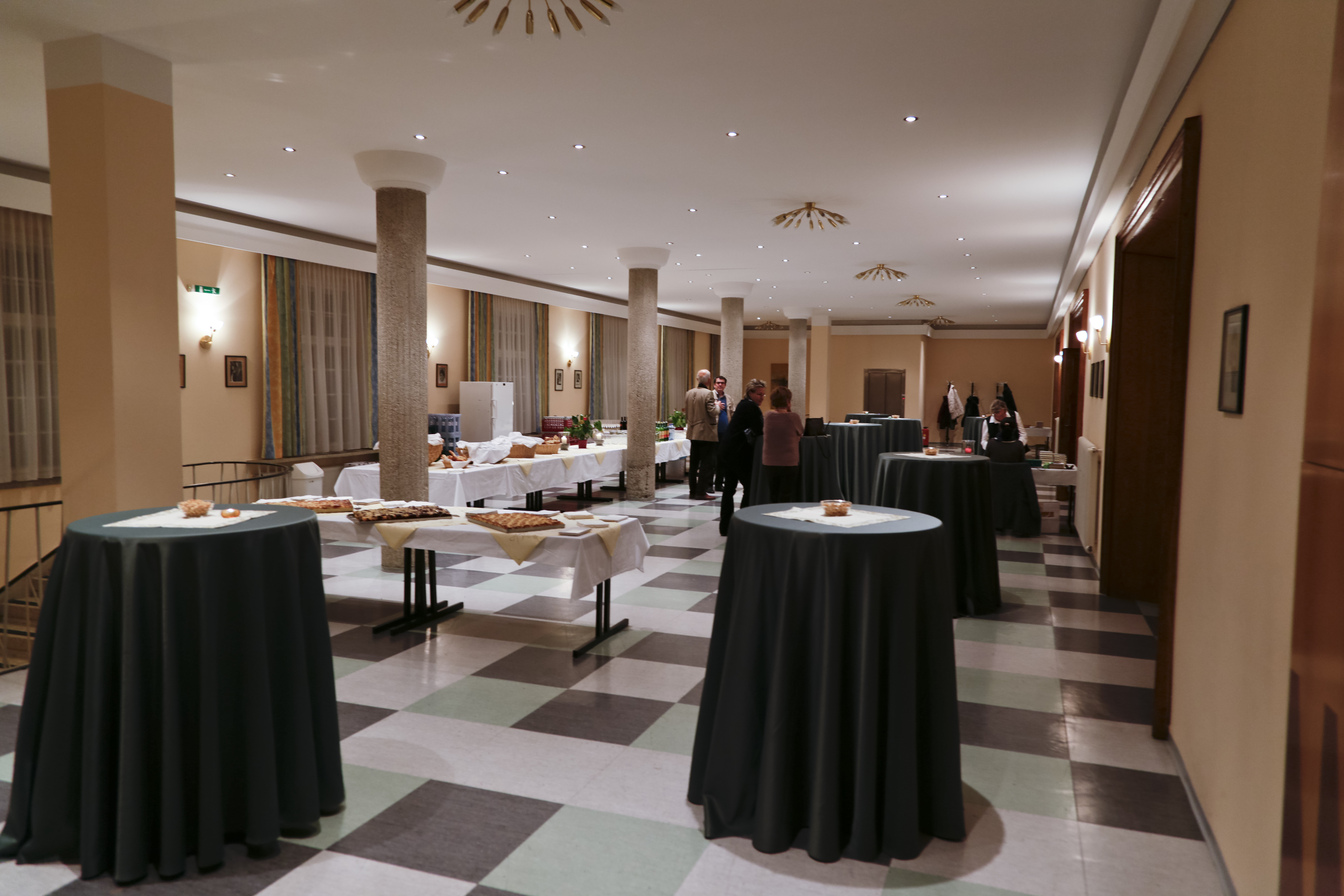 Ein offener Raum mit Buffet und Stehtischen. Mehrere Säulen, Wand- und Deckenleuchten schaffen ein warmes abendliches Ambiente.