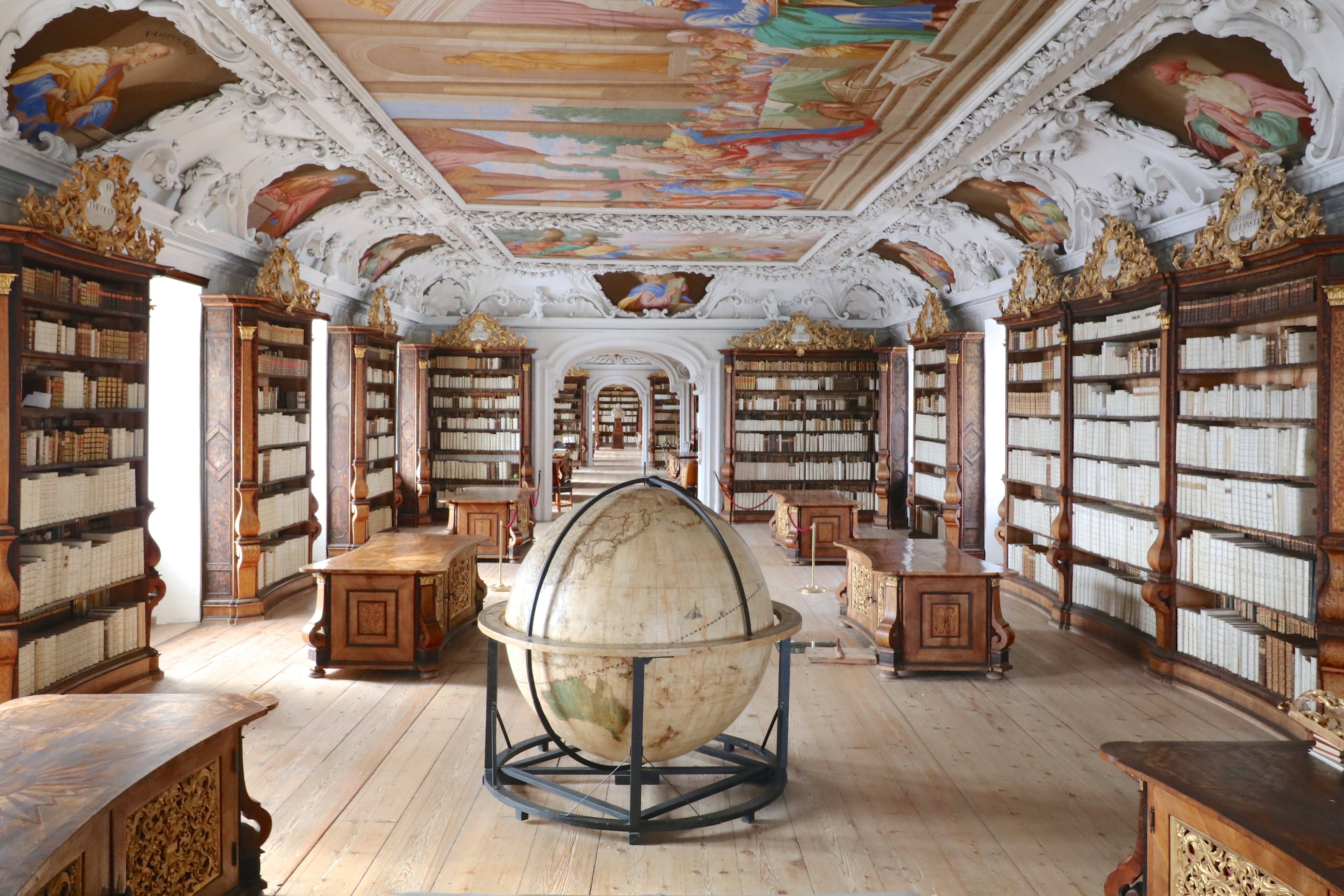 Eine große, prunkvolle Bibliothek mit hochwertigen Holzschränken und Goldverzierungen; auch die Decke ist verschnörkelt und mit Gemälden bemalt