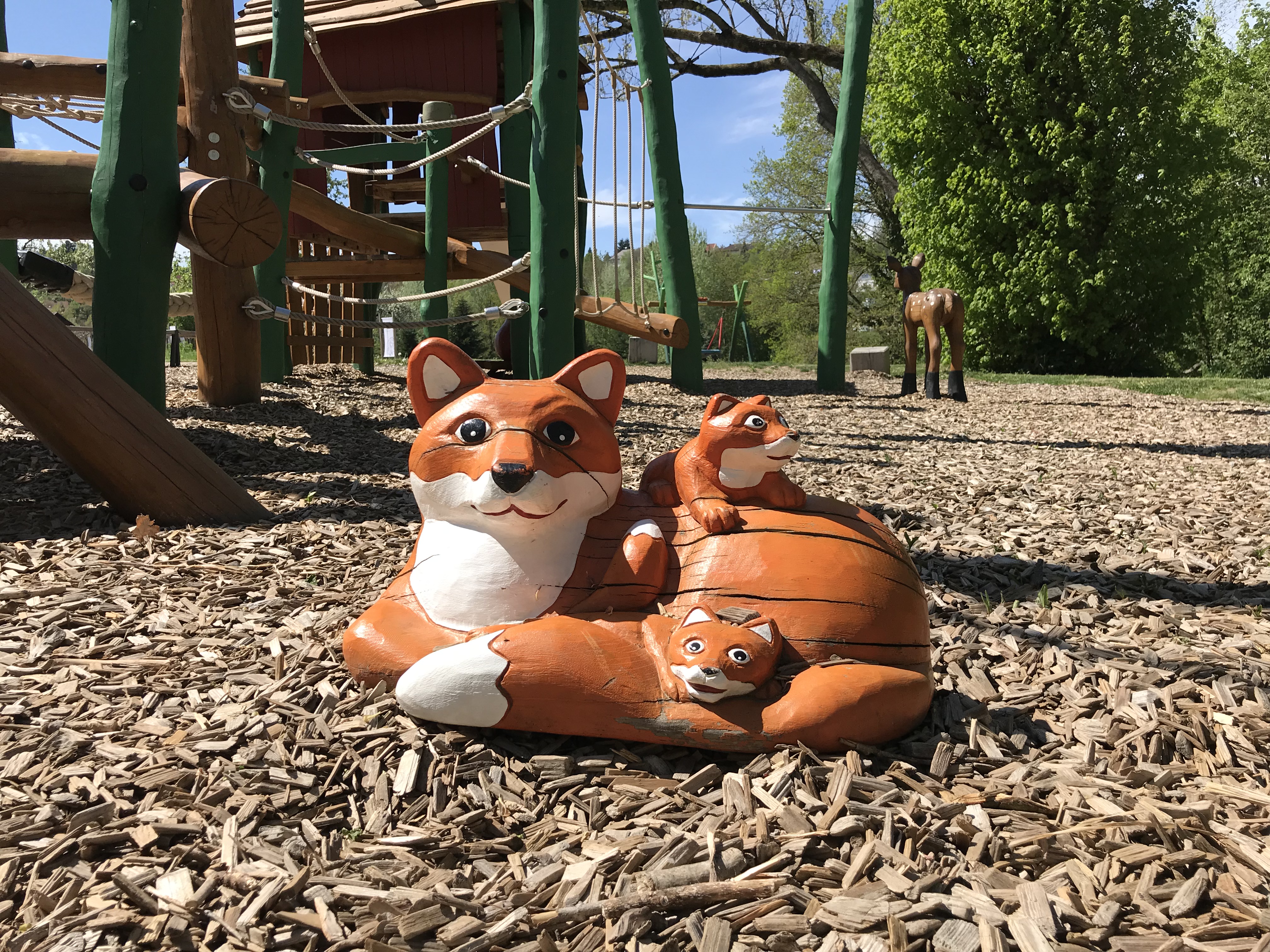 Eine hölzerne Fuchsmutter liegt mit ihren zwei Kleinen am Spielplatz und lächelt