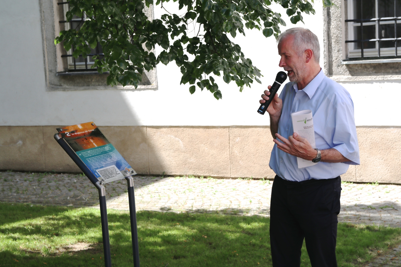 Ein Mann liest im grünen Stiftshof von einer Informationstafel und spricht ins Mikrofon