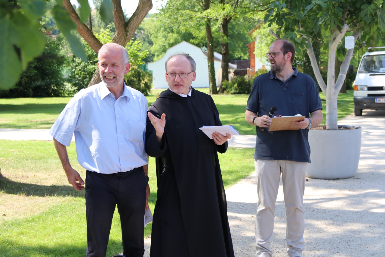 Ein Mönch weist einem Mann per Handbewegung den Weg. Die beiden und ein Mann dahinter befinden sich auf einem Kiesweg der Stiftsgärten.