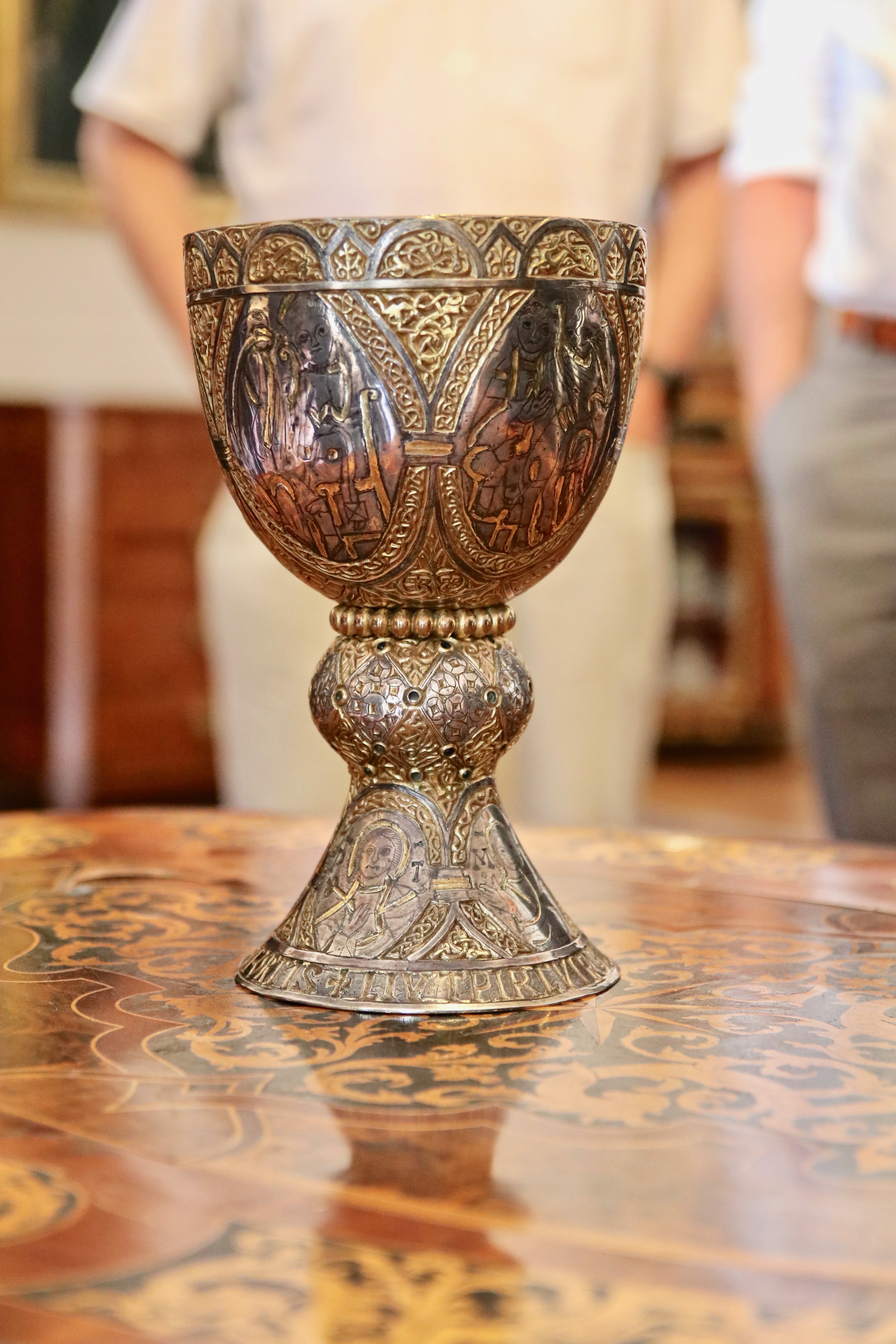 Nahaufnahme des Tassilo-Liutpirc-Kelches: silber- und goldfarbene Edelmetalle bilden einen robusten Kelch, der durch Inschriften, Abbildungen von Heiligen und einem Perlenkranz beeindruckt