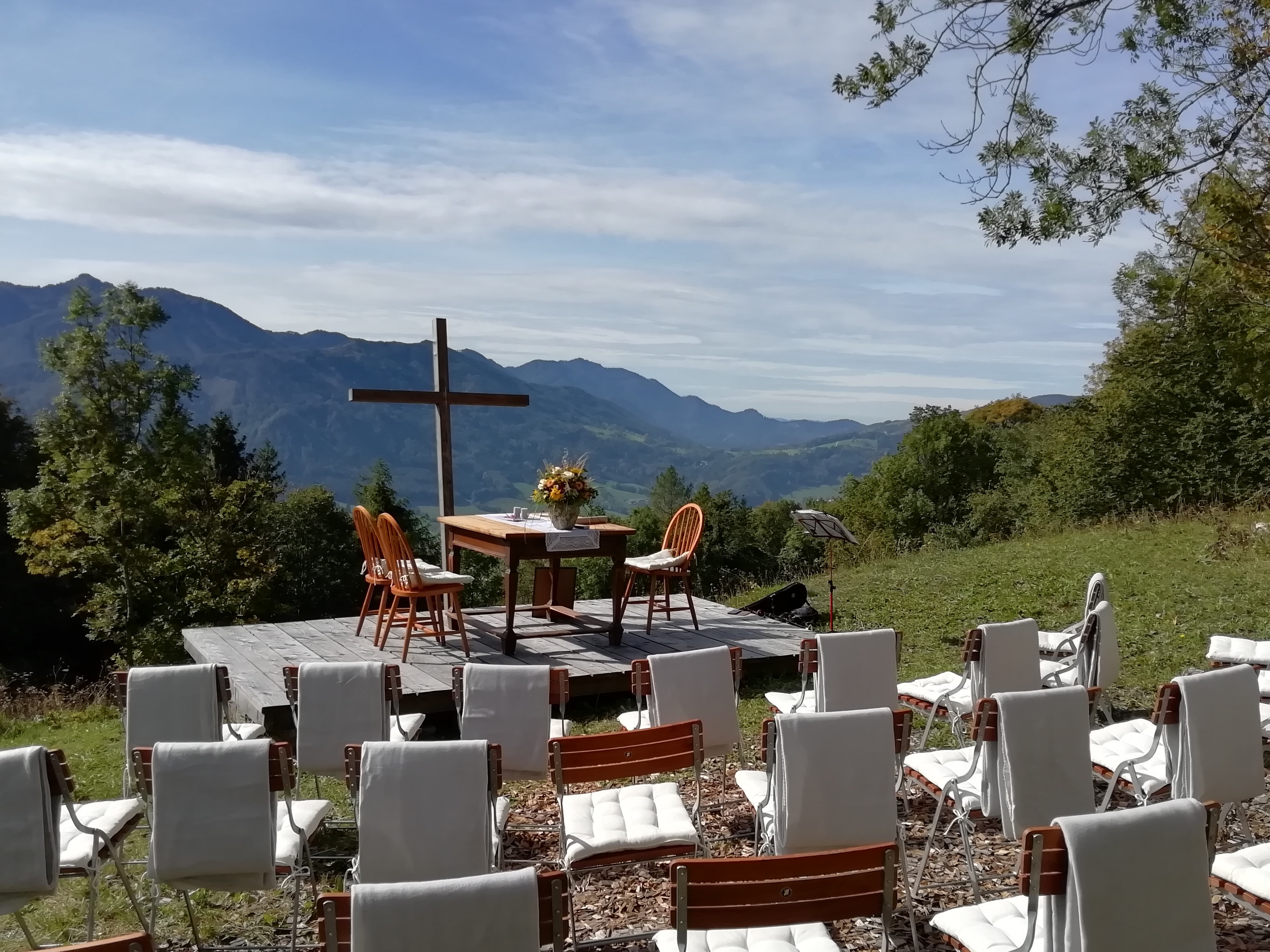 Eine Bestuhlung zur Trauung mit Blick auf ein hölzernes Jesuskreuz. Im Hintergrund eine mit Wald bedeckte Hügellandschaft.