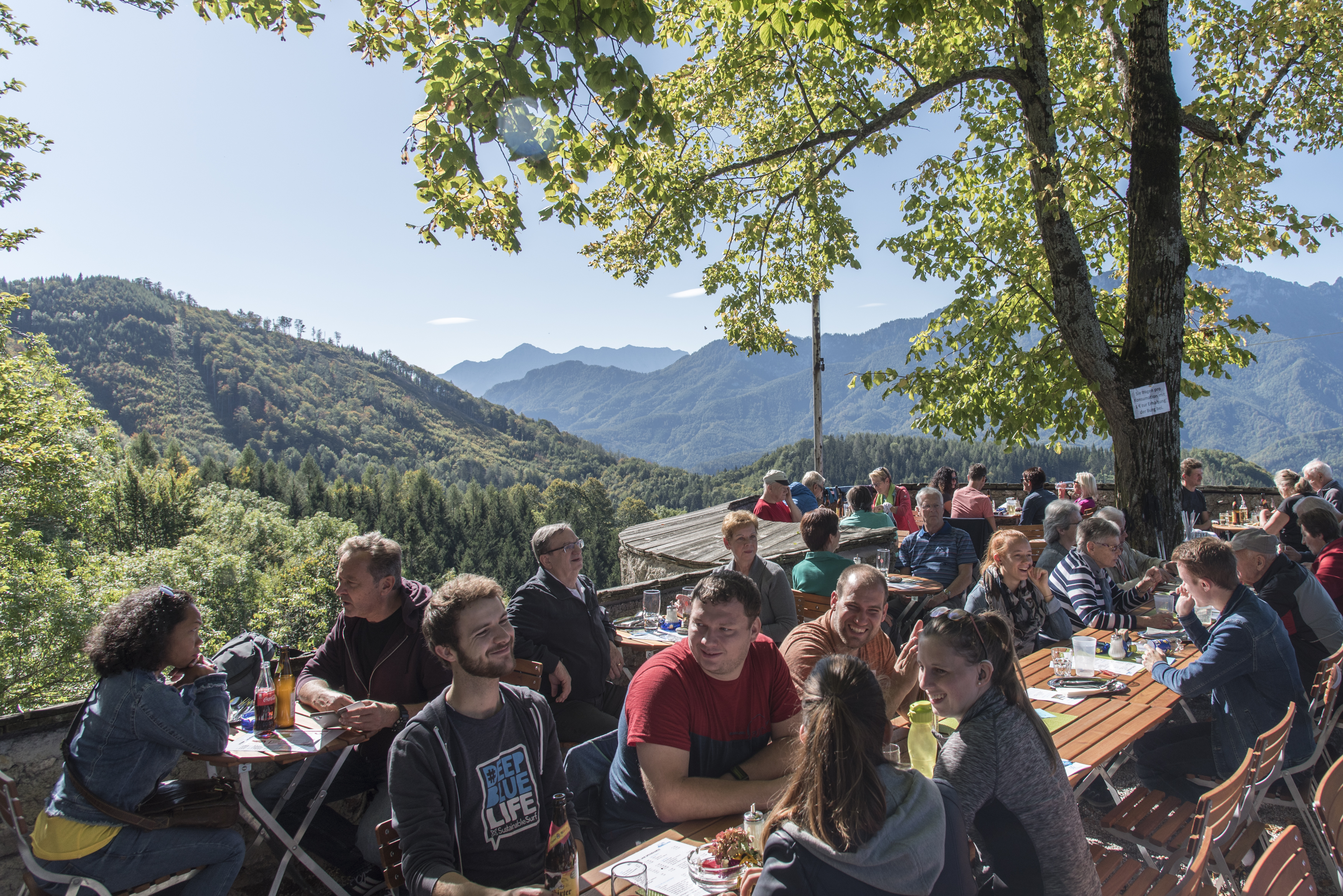 Auf der Burgterrasse sitzen viele Menschen gemütlich beisammen. Ein Baum spendet Schatten. Im Hintergrund eine mit Wald bedeckte Hügellandschaft.