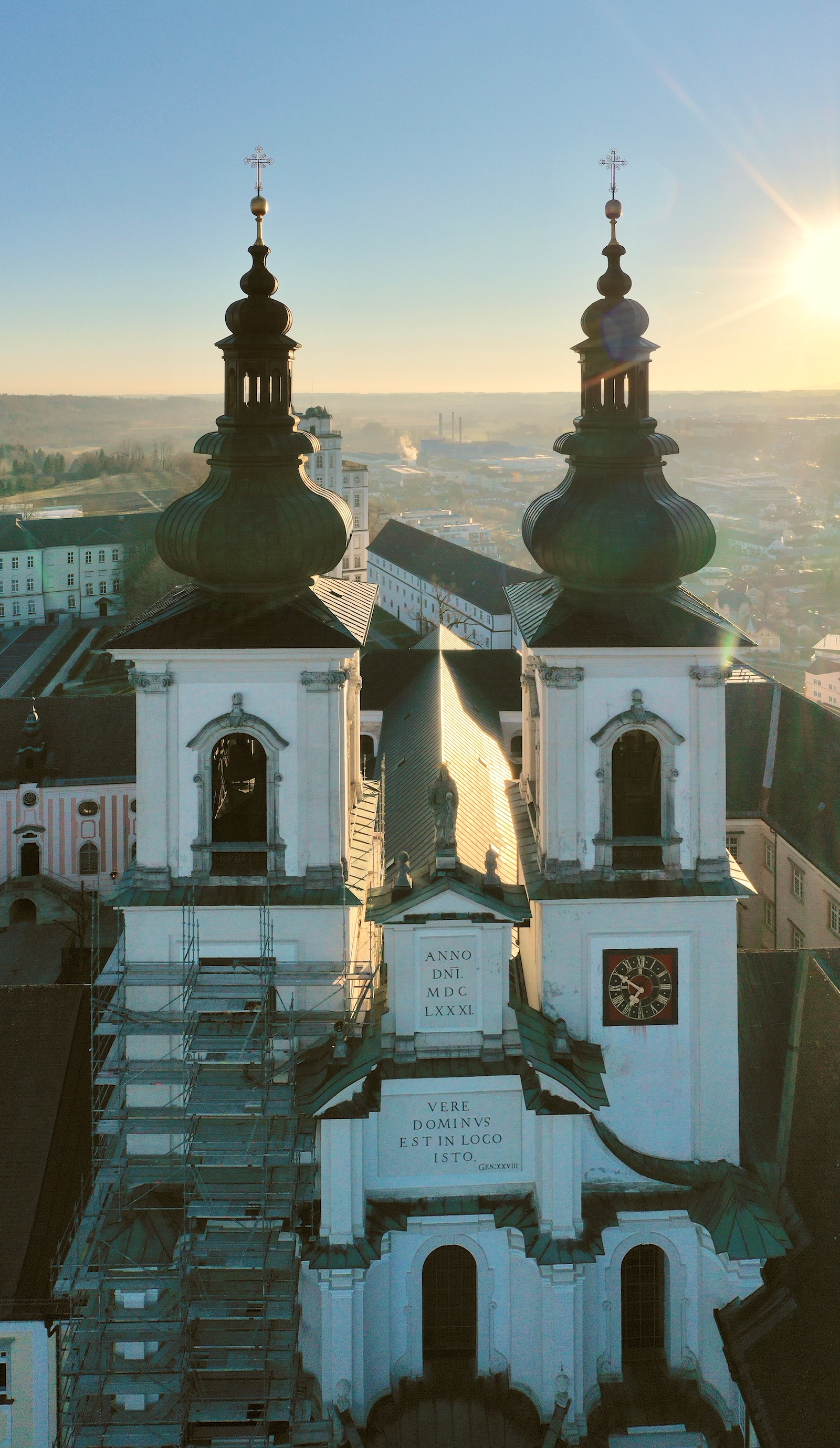 Vogelperspektive auf die Stiftskirche; der linke Kirchenturm ist mit einem Gerüst verkleidet; im Hintergrund steigt die Sonne auf, es ist Vormittag