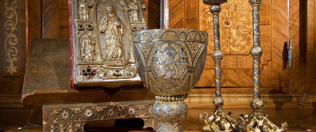 Buch, Kelch und Leuchter aus edlen Metallen gefertigt und mit Jesus Christus und verschiedenen Heiligen verziert. Das Buch enthält mehrere rote Edelsteine am Umschlag, sowie einen roten Buchrücken aus Leder.