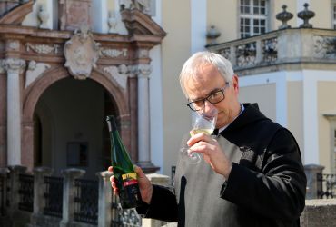 Ein Mönch steht mit einer Flasche Wein und einem Weinglas vor dem Haupteingang und verkostet den aktuellen Jahrgang