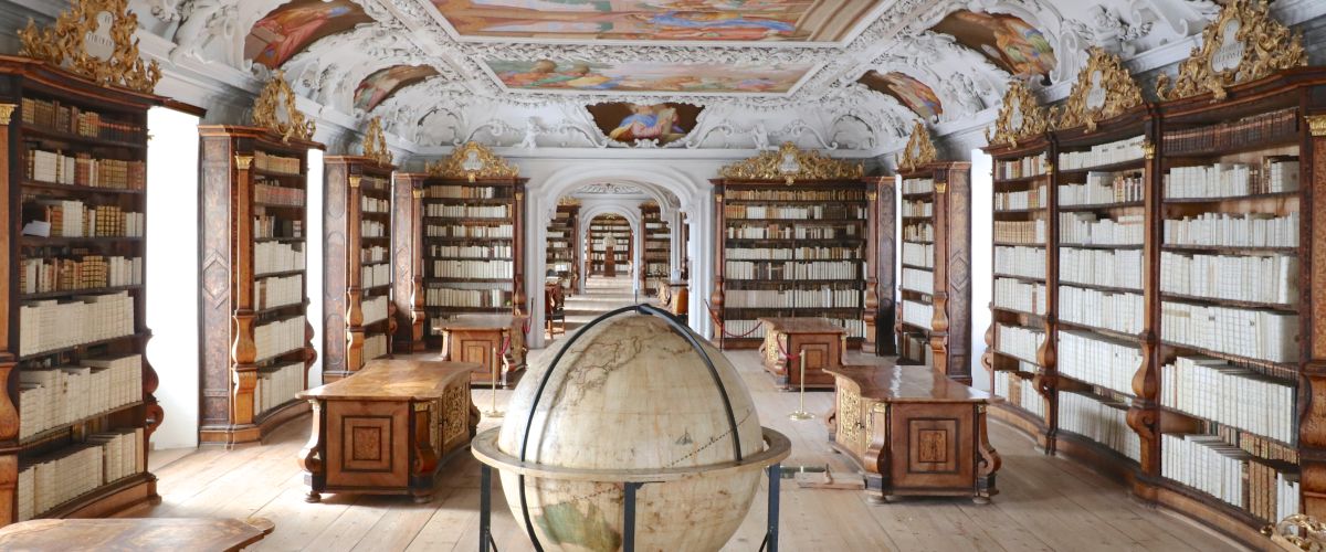 Eine große, prunkvolle Bibliothek mit hochwertigen Holzschränken und Goldverzierungen; auch die Decke ist verschnörkelt und mit Gemälden bemalt