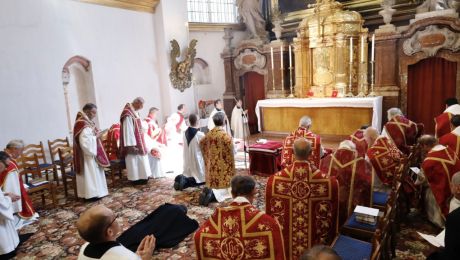 Priester in rotem Messgewand vor Altar, in der Mitte liegt ein Mönch ausgestreckt auf dem Boden