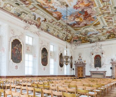 großer barocker Festsaal mit Deckenfresko und Stuck an den Wänden, mit aufgestellten Stühlen
