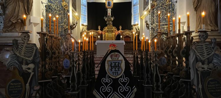 ein in schwarz gehüllter Sarg, umgeben von kerzentragenden Skeletten aus Holz mit blick auf altarbild schwarz verhangen