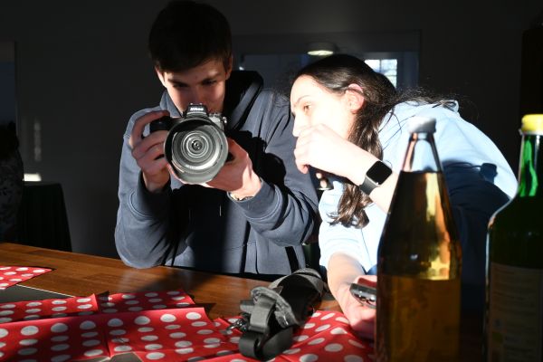 Ein junger Mann fotografiert mit seiner Kamera direkt in die Fotoaufnahme. Eine junge Frau blickt interessiert auf s eine Kamera und wird dabei stark von der Sonne erhellt.
