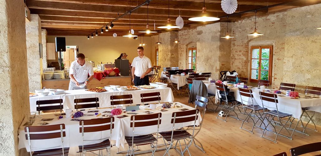 Zwei Kellner decken die Tische zwischen Mauern aus Stein und einer warmen, einladenden Holzdecke. Eine Lichtleiste an der Decke schafft gemütliches Ambiente.