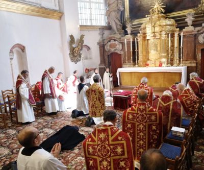 Priester in rotem Messgewand vor Altar, in der Mitte liegt ein Mönch ausgestreckt auf dem Boden
