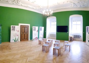 Ein Raum mit Parkettboden, frisch-grünen Wänden und einer weißen verschnörkelten Decke. Moderne Holzhocker bieten Platz beim Blick auf den Flat-TV