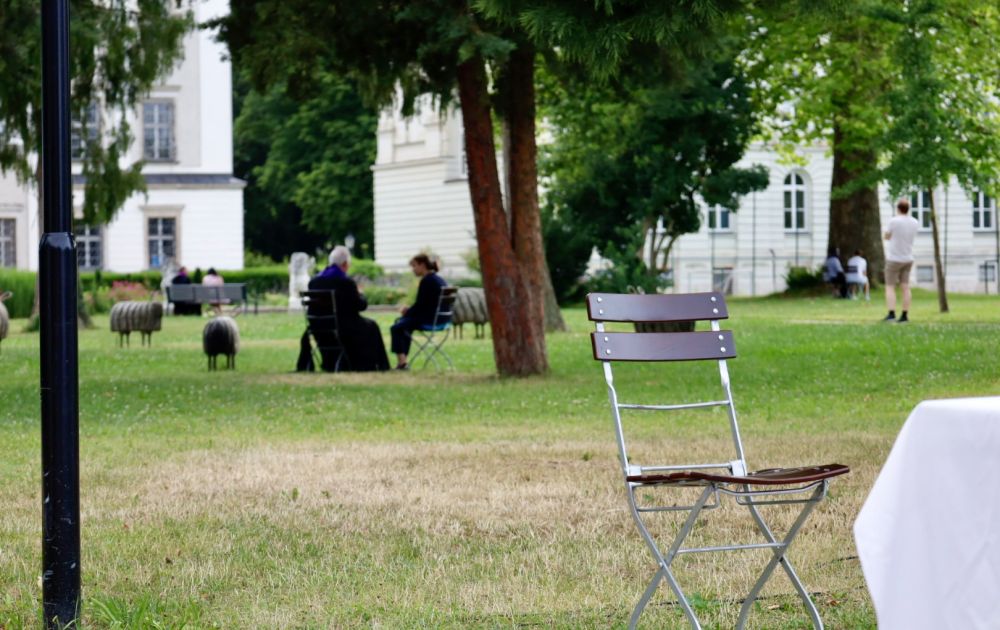 Auf der Wiese der Stiftsgärten werden vereinzelt Beichtgespräche abgehalten. Ein leerer Stuhl im Bildvordergrund lädt zur Seelsorge ein.