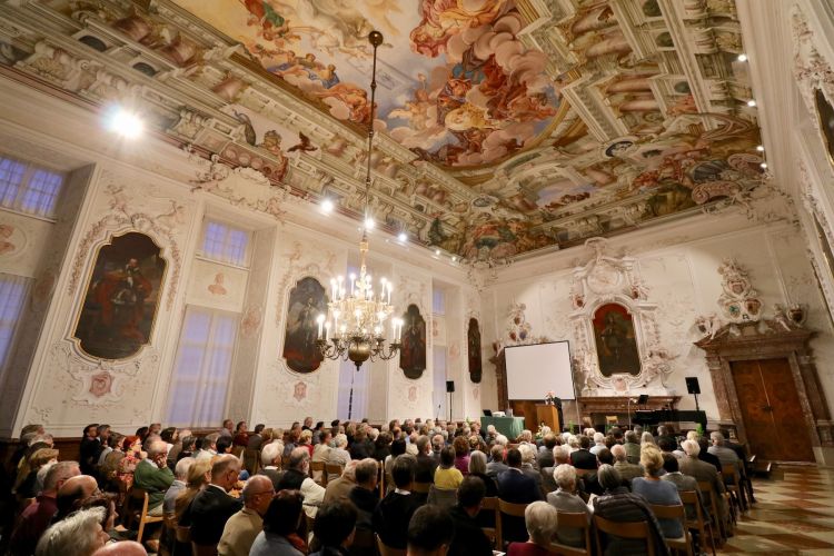 Ein prunkvoller Saal mit vielen Verschnörkelungen und Gemälden an den Wänden und der Decke, gro0e Kronleuchter und Schweinwerfer geben Licht für das große Publikum und den Referenten