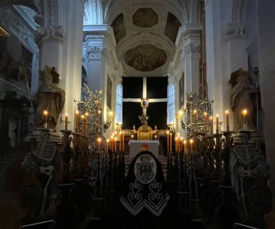 ein in schwarz gehüllter Sarg, umgeben von kerzentragenden Skeletten aus Holz mit blick auf altarbild schwarz verhangen