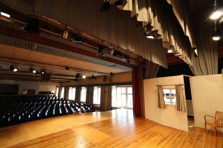 Blick von der hölzernen Bühne im Theatersaal. Die Scheinwerfer auf der Bühne sind an. Hunderte Sitzplätze stehen leer im dunklen und warten auf die nächste Theater-Aufführung.