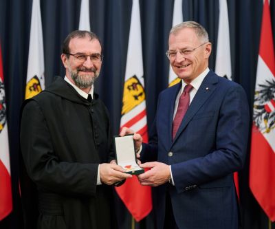 Zwei Personen, der Landeshauptmann übergibt die Verdienstmedaille an den Geehrten, in Hintergrund oberösterreische Fahnen