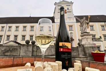 Eine Flasche Wein und ein Achterl Wein stehen auf einem Holzfass, dekoriert mit vielen Korken, im Hintergrund die Fassade eines Stiftsgebäudes