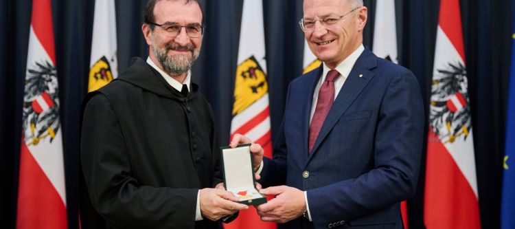 Zwei Personen, der Landeshauptmann übergibt die Verdienstmedaille an den Geehrten, in Hintergrund oberösterreische Fahnen
