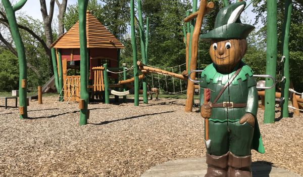 Eine grüne Holzfigur stellt Robin Hood dar. Sie lädt zum Klettern am dahinter liegenden Kinderspielplatz ein. Der Boden ist mit Holzsägespänen bedeckt.