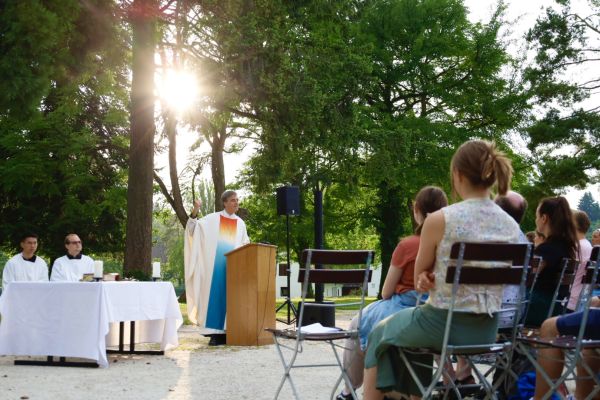 Pater Bernhard predigt im freien beim Sommerfest zur Menge, im Hintergrund scheint die Sonne durch die Bäume der Stiftsgärten