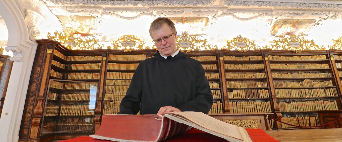 Mönch blickt in ein großes, dickes Buch; dahinter türmen sich große Bücherregale; die Decke ist hell beleuchtet