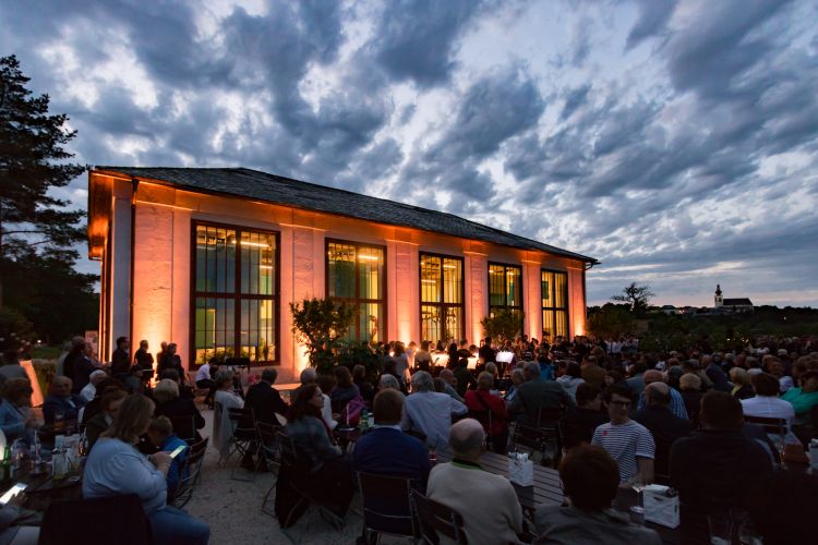Große Veranstaltung vor dem Feigenhaus unter freiem Himmel. Die Nacht bricht ein und das Feigenhaus mit dessen großen Fenster und Blumenstöcke erstrahlt in dekorativer Beleuchtung