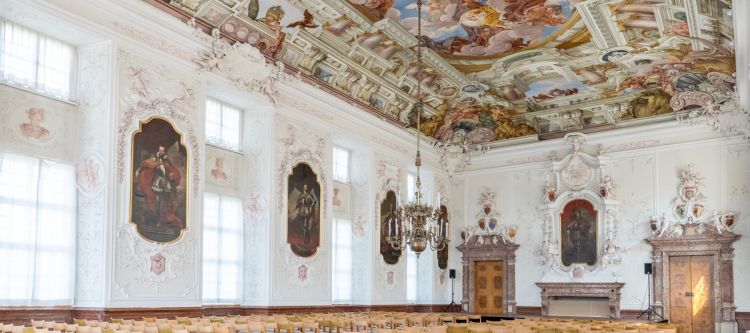 großer barocker Festsaal mit Deckenfresko und Stuck an den Wänden, mit aufgestellten Stühlen