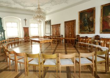 Viele leere Sessel bilden einen Kreis. In der Mitte ein Holztisch im Jugendstil. An der Wand ein hölzernes Jesuskreuz.