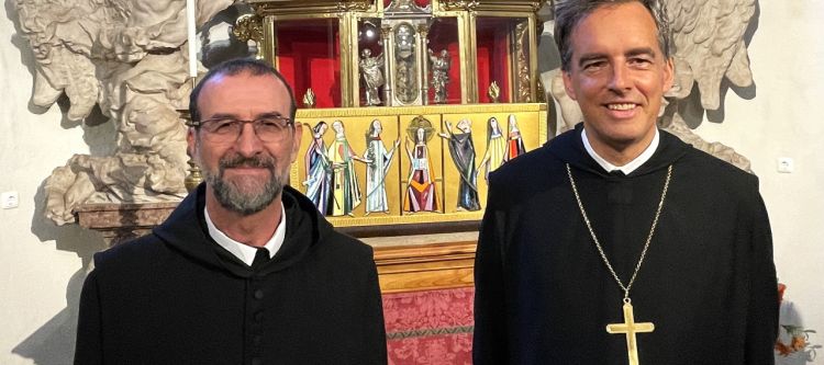 Mönch in schwarzem Ordengewand spricht in Mikrofon vor Priestern in rotem Messgewand