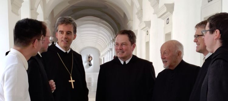 Mönche in schwarzem Ordengewand begrüßen einen Gast, im Hintergrund ein langer Gang