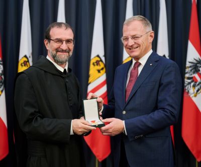 Zwei Personen, der Landeshauptmann übergibt die Verdienstmedaille an den Geehrten, in Hintergrund oberösterreische Fahnen
