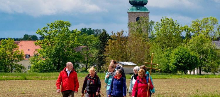 Kleine Pilgergruppe auf Wiesenweg mit Kirche im Hintergrund