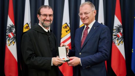 Zwei Personen, der Landeshauptmann übergibt die Verdienstmedaille an den Geehrten, in Hintergrund oberösterreische Fahnen