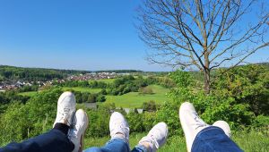 Die Beine dreier Jugendlicher sind hochgelegt: Jeans und weiße Sneaker zeigen Richtung satter Hügellandschaft