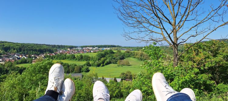 Die Beine dreier Jugendlicher sind hochgelegt: Jeans und weiße Sneaker zeigen Richtung satter Hügellandschaft