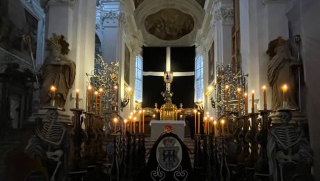 ein in schwarz gehüllter Sarg, umgeben von kerzentragenden Skeletten aus Holz mit blick auf altarbild schwarz verhangen