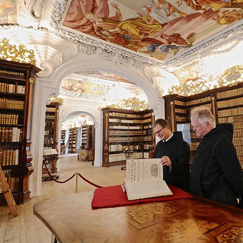 Zwei Mönche stehen an einem Tisch in der Bibliothek und blicken in ein großes, dickes Buch