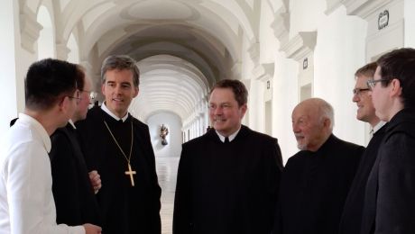 Mönche in schwarzem Ordengewand begrüßen einen Gast, im Hintergrund ein langer Gang